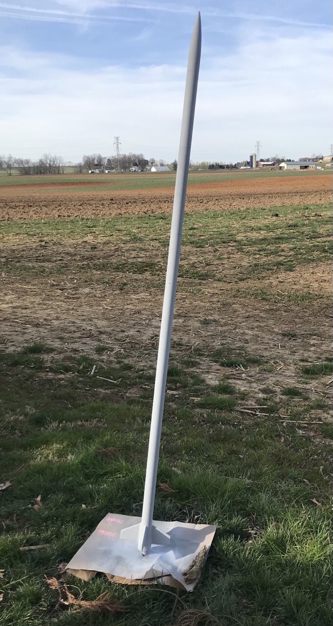 A model rocket standing next to field with a fresh coat of primer, looking very much like the profile image for this account.