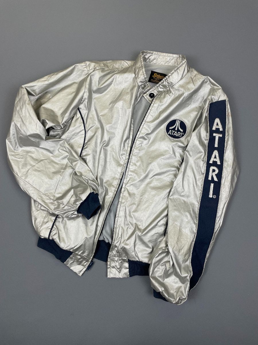 A vintage silver satin bomber jacket with navy blue ribbed cuffs, collar, and waistband, featuring the classic Atari logo patch on the left chest and bold “ATARI” lettering running vertically down the right sleeve. The jacket is laid flat on a gray background, zipper partially open.